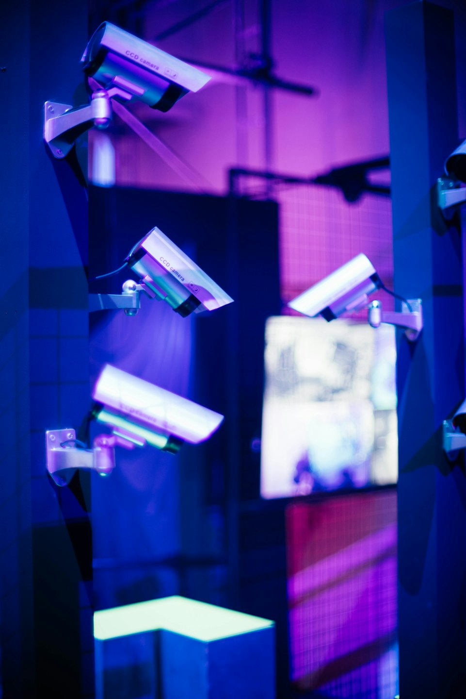 Multiple grey-white security cameras on blue walls with purple lighting.